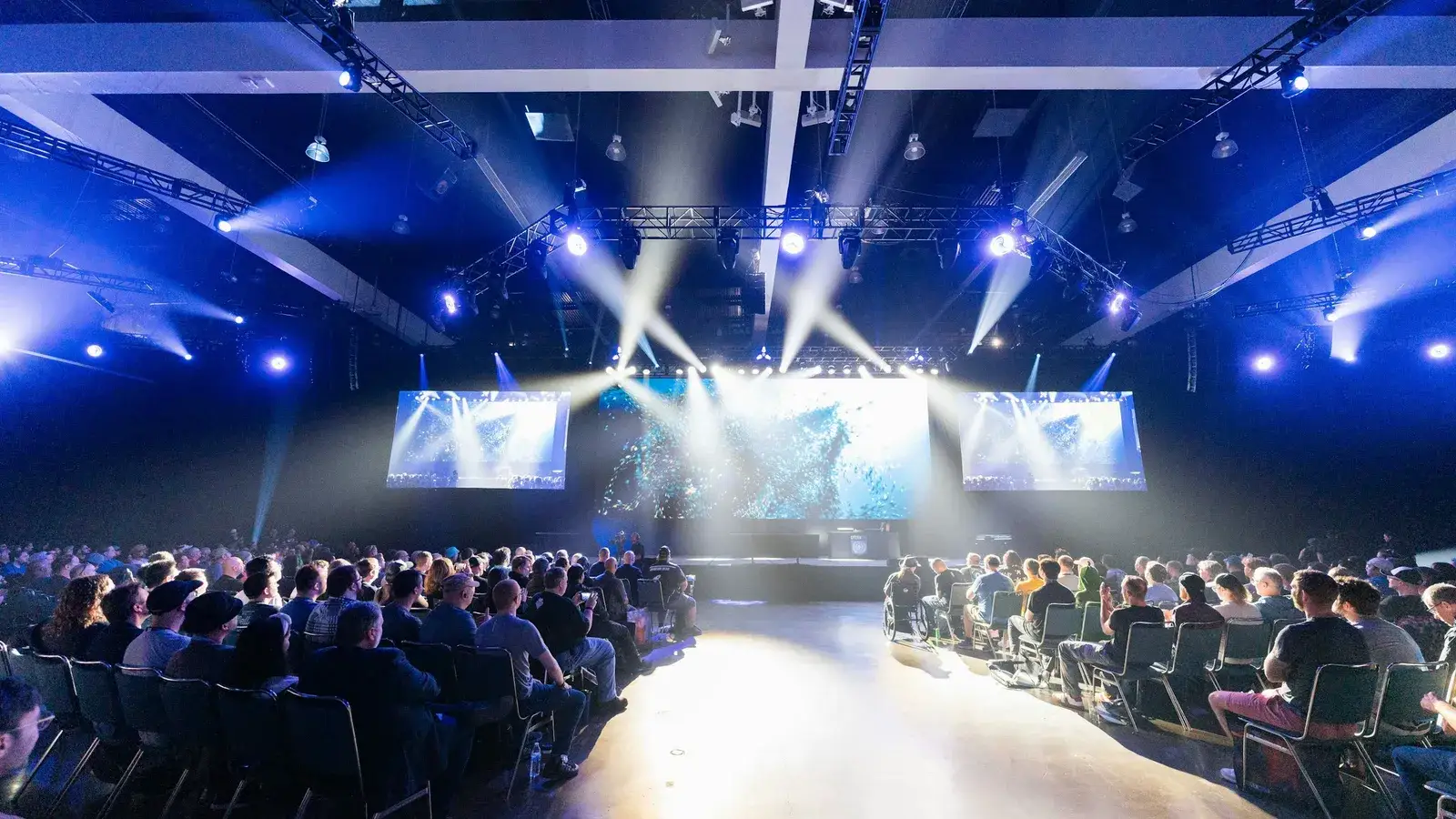 An audience in a large hall under bright blue stage lights