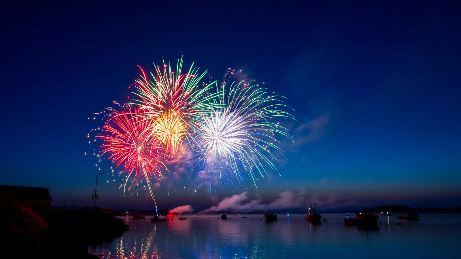 Fireworks bursting over water at dusk, reflected in the lake below